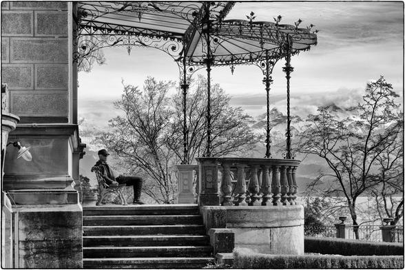 Schwarzweissfotografie einer Loggia mit einer Bank, worauf ein Mann sitzt. Im Hintergrund sind Berge und ein See zu sehen.