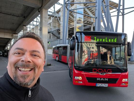 Marcus steht am Rand der Strasse auf der man sieht, wie ein roter Gelenkbus mit der Beschriftung "DOAG JavaLand" das Gelände des Nürburgrings verlässt.