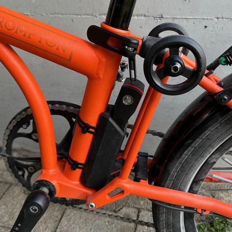 orangenes Faltrad im aufgeklappten Zustand. Das Fahrradschloss befindet sich zwischen Sattelrohr und Hinterbau. Alles sitzt, wackelt und hat Luft - als wäre es dafür gemacht.