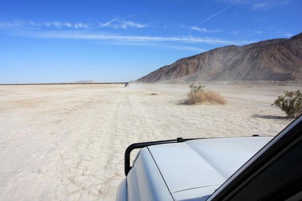 Two Toyota Land Cruisers zip along the flat desert, with abrupt hills on the right in the background. The view is from a GoPro camera mounted somewhere above the driver side window on the rear Land Cruiser.