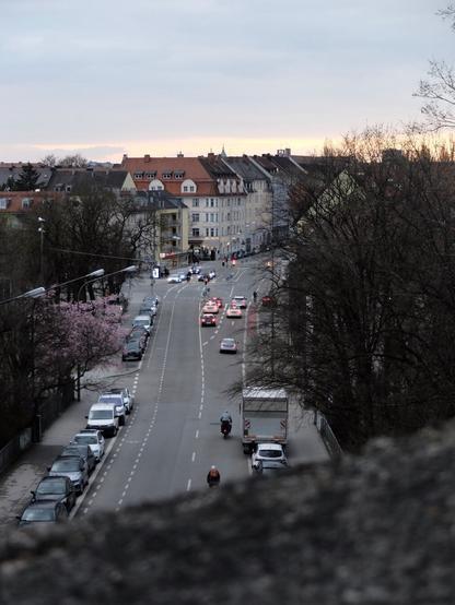 A city street view from an elevated perspective at dusk. The road is lined with parked cars on both sides, with a bike lane on the right. A few cyclists and vehicles are moving along the street, illuminated by streetlights and car headlights. Residential buildings with red-tiled roofs line the street, and blooming cherry trees are visible on the left. The sky is overcast with a hint of sunset on the horizon. Bare trees frame the right side of the image.