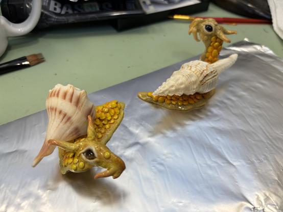 Two yellow-orange painted "dragon-snails" crafted out of epoxy clay, set into real seashells.  The one in front has a long puppy-dog face, while the one in back is perked up curiously.