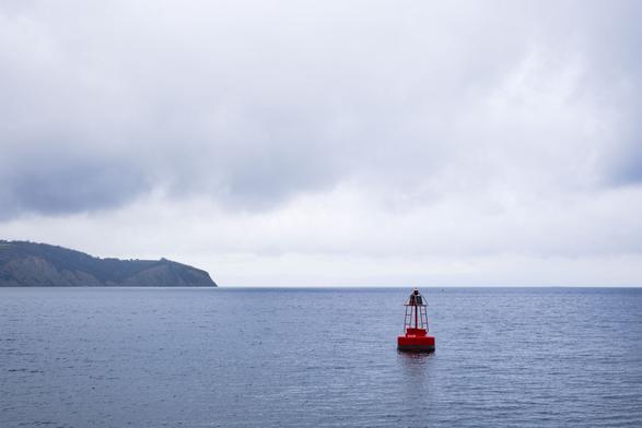 Sea and a red buoy on the right. At the back left you can see part of a headland. The sky is slightly cloudy and takes up the upper two thirds of the photo.