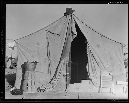 The image depicts a dilapidated tent structure with large openings and sagging fabric, suggesting it is weathered or in disrepair. A single hat hangs from the peak of the tent's entrance. In front of the tent are various objects: an old-fashioned metal tub possibly used for cooking or washing; another cylindrical container that appears aged; a small bucket on the ground with its contents not visible due to lighting and shadowing effects; a wooden plank leaning against what could be part of an interior wall, serving as a makeshift step leading into the tent. The scene is set in daylight under clear skies, evidenced by strong shadows cast across the objects and floor.

The photograph conveys a sense of abandonment or hardship with its focus on the worn-out appearance of the tent and sparse surroundings. There's no direct indication of human presence within this frame; however, one can infer that it may belong to someone who lives in temporary quarters due to conditions suggested by the setting. The image has an historical feel likely dating back several decades based on the style of objects depicted—such as the metal tub—and overall composition and black-and-white tonality.

This photo is part of a larger collection, potentially documenting life during times of economic hardship or displacement, such as in rural agricultural communities where workers like pea pickers might reside temporarily. The mention of Calipatria sugg [...]