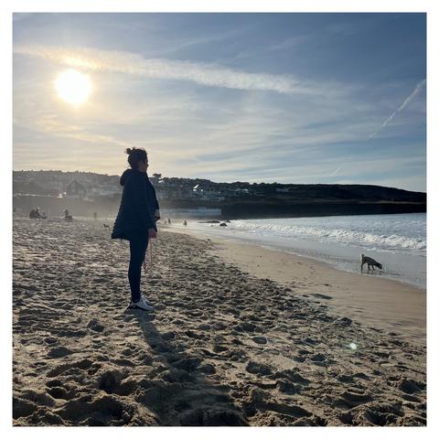 Beach scene, late in the day sun low in the sky a silhouetted female figure stands frame left looking out to sea with a small dog (far frame right) searching in the incoming tide.