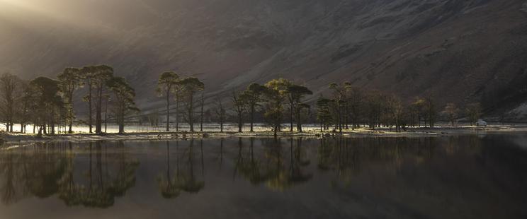 A line of pine trees line the banks of a lake with a small white painted cottage at the far end. Behind are the lower slopes of a mountain (Haystacks). The trees are reflected in the perfectly still waters. Sunlight breaks through a gap in the mountains to backlight the trees.
