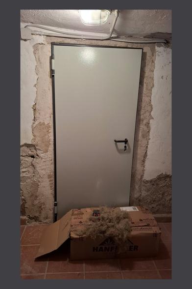 The image shows a metal door installed in a rustic, unfinished wall. The surrounding wall has exposed plaster and areas of rough, uneven texture, indicating ongoing renovation work. A light fixture is mounted on the ceiling, with electrical conduit running along the top.
In front of the door, there is an open cardboard box labeled "HANF" (likely referring to hemp insulation material). There are two spatulas on the box. Some loose hemp fibers are spilling out of the box onto the tiled floor. The scene suggests that insulation work may be in progress, possibly for sealing gaps around the door or improving thermal efficiency in the space.