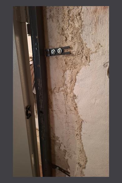 This image shows a close-up of the door frame and its mounting structure. The door is slightly open, revealing the metal frame and the wall behind it. The wall has a rough plastered surface with an uneven texture, indicating prior renovation work.
Two black metal brackets are secured to the wall with screws, helping to anchor the door frame. The brackets have an open slot design, possibly to allow for adjustment before final tightening. The frame itself appears to be a sturdy metal construction with ventilation or drainage slots.
This view gives insight into how the door was fixed into the wall, showing both the structural integration and the unfinished nature of the surrounding plasterwork.