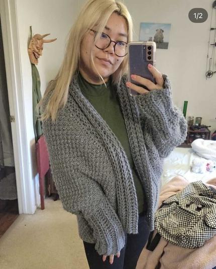A Japanese woman with blonde medium long length hair wearing a green shirt, black pants and a handmade grey cardigan wrap. The cardigan is knit with a bulky weight yarn and is held double with a silver tinsel yarn (although the tinsel is not visible in the photo). The woman is posing with her head cocked slightly to her left. She has a phone in one hand with the other hand resting on her leg. Her bedroom is in the background.