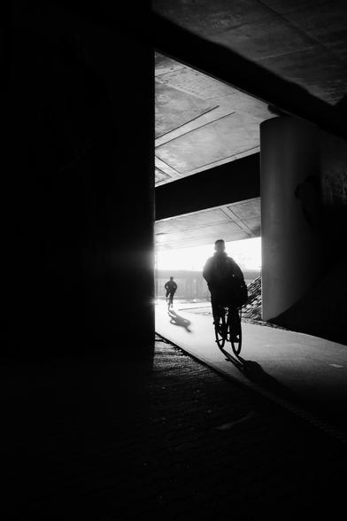 backlight high contrast B&W photograph two cyclists a shilouets, highway underpass, lineplay from ceiling and bikeway, dark shadows