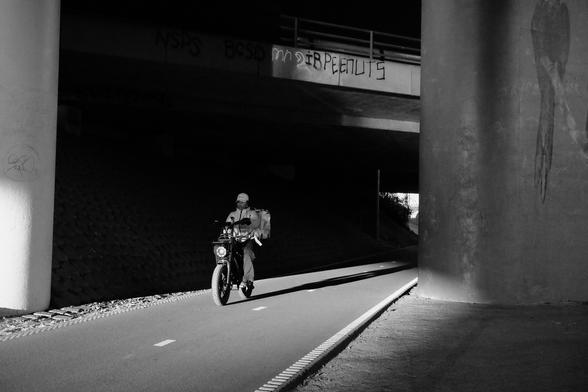 B&W photograph, low sun evening shot, cyclist courier coming out of underpass of highway,high contrast, dark shadows.
