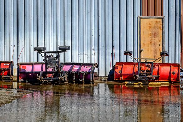 A pink and orange snowplow sit at the edge of a large puddle in front of a metal sided building.
