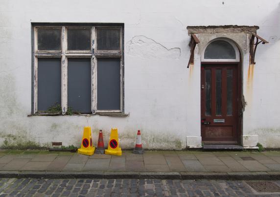 A straight on photo of the front of a white painted building, to the left is a 6 pane window where the lower pains are painted grey and the white painted woodwork is showing the grey undercoat and missing rotten wood. Rust streaks stain the wall from brackets that once held a now missing roof over the door. There is a selection of traffic cones against the green stained lower section of the wall.