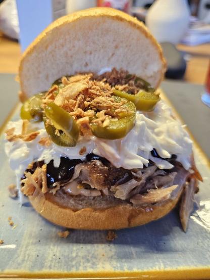 Das Bild zeigt einen appetitlich aussehenden Sandwich, der auf einem blauen Teller mit gelbem Rand serviert wird. Der Sandwich besteht aus einem weichen, braunen Brot, das halb geöffnet ist, um die Füllung zu zeigen. Die Füllung besteht aus zerkleinertem, braunem Fleisch, das wahrscheinlich Rindfleisch ist, und ist mit einer dunklen, glänzenden Sauce bedeckt, die möglicherweise eine Art Barbecue-Sauce ist. Auf dem Fleisch befindet sich eine Schicht weißer, cremiger Sauce, die wie eine Art Ranch-Dressing aussieht. Darüber sind grüne Jalapeño-Scheiben und goldbraune Kräuterflocken, vermutlich Frittierte Zwiebeln, arrangiert. Der Hintergrund ist leicht unscharf, wodurch der Fokus auf dem Sandwich liegt

Bereitgestellt von @altbot, privat und lokal generiert mit Ovis2-8B