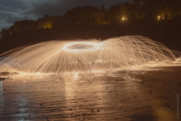 A long-exposure photograph of steel wool spinning on a beach at night, creating a dramatic effect of golden sparks radiating outward in circular patterns. The dark silhouette of trees and faint lights in the background contrast with the fiery display.
