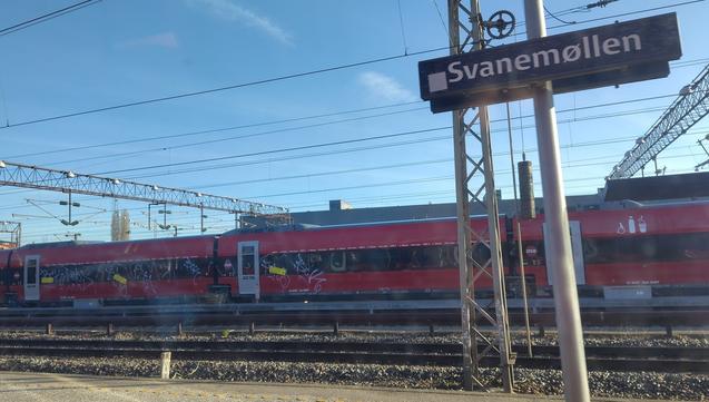 Modern-looking train cars in DSB red partially covered in grafitti. Station sign: Svanemøllen.
