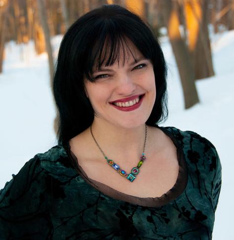 Catherynne Valente, a white woman with long black hair, wearing a dark green top and a necklace with large multicolored beads, standing in snowy woods.