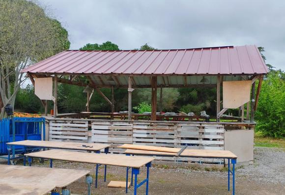 Photo d’une cabane un peu tristounette en bois de palettes et un toit en fer devant sont entreposés des table de type  « mairie »