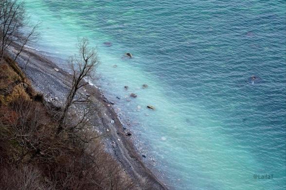 Blick einen Steilhang hinunter zum Strand und zur See. Der Steilhang ist mit kahlen Bäumen und Büschen bewachsen, der Strand ist ein recht schmaler, steiniger, geschwungener Streifen, und die See zeigt sich in verschiedenen Türkistönen. Zum Strand hin ragen Steine aus dem Wasser.