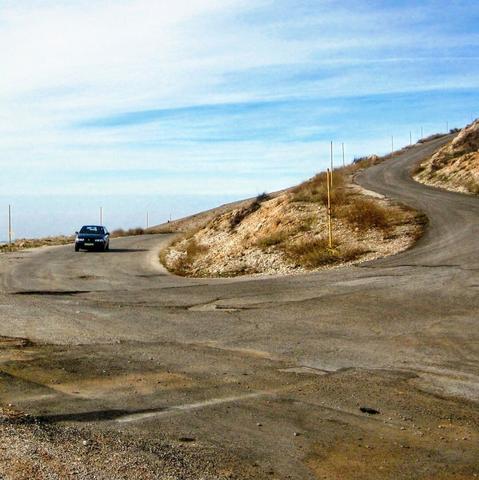 A wide asphalt road leads up a mountain in curves. A single car drives the way up.  blue sky with white clouds