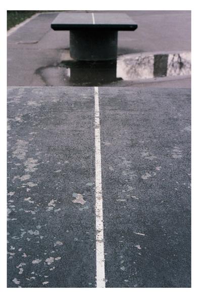 Color photograph of a table tennis table in Donaupark, Vienna. The close-up shows that the table tennis table is quite weathered. The surface has cracks. The view wanders over the table to a second table tennis table. This one also has no net and is worn out. A puddle of rain can be seen between them.