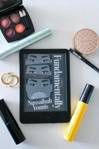 Kindle version of novel Fundamentally by Nussaibah Younis. Book cover features gradually expanding view of woman: first just striking eyes and eyebrows, then a little more, and finally same woman blowing a bubble. Kindle is surrounded by various makeup.