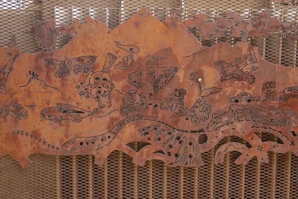 Orange iron panel covers a decorative grill for the back of seating at an outdoor cafe.  The quest or battles of this decorative iron "drawing" seem to have different interpretations depending on what is going on in your world when you see it.  Maybe  Day of the Dead skeletons trying to outrun attacks.  But then I wonder if the helmets and surreal images show Nazi villains.  Intricate surreal drawing and unusual rusted metal panel.