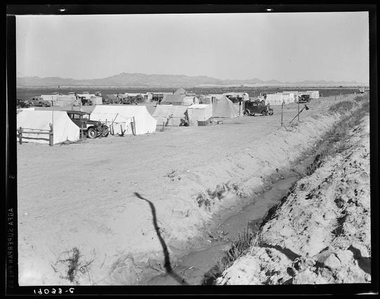 The image depicts a rural landscape with rows of white tents and makeshift structures scattered across the sandy terrain. The scene is reminiscent of temporary accommodations for agricultural workers, possibly in California during the late 19th or early 20th century. Old-fashioned cars are parked among the buildings, adding to the historical ambiance.

In the foreground, a dirt road meanders through the campsite with tire tracks visible on its surface. A shallow ditch runs parallel to one side of the path and contains some stagnant water that reflects part of the landscape above it. The background is dominated by rolling hills or mountains under an overcast sky, which contributes to the overall rustic setting.

The black-and-white composition suggests this photograph was taken during a time when color photography wasn't widely accessible or used for everyday documentation purposes.