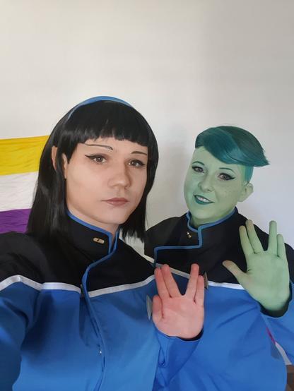 Two cosplayers dressed as the characters T'Lyn and Tendi from the TV show Star Trek: Lower Decks. They both do the Vulcan salute towards the camera. In the background is a nonbinary pride flag.