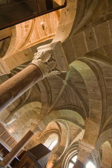 Inside of sandstone (red and blonde) hall, supported by columns. Carved capitals, round romanesque arches. The photo is strongly slanted, worm’s-eye view.