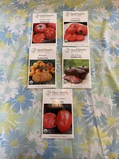 Five rectangular seed packs on a floral sheet. These are Seed Savers Exchange varieties, Eva Purple Ball, Amish Paste, Moonglow, Paul Robeson, and Salvaterra’s Select