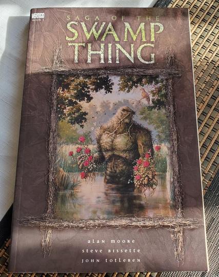 Comic book cover. Dominant brown background with vines. Centered composition showcases an illustration of Swamp Thing, a humanoid plant creature, framed within a vine border against a swamp scene with foliage, a bird, and roses in its hands. "Saga of the Swamp Thing" is in textured stone white text. Vertigo logo and "Book 1" in the upper left. The names "Alan Moore, Steve Bissette, and John Totleben" are typeset in white near the bottom, arranged vertically.