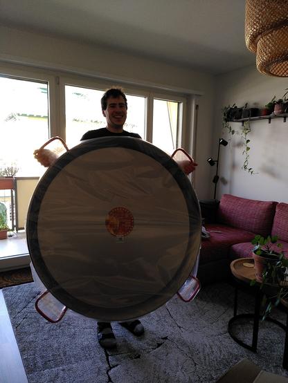 Me standung in my living room holding up a hige Paella pan. At a diameter of 1.15m, the pan is so big that it hides most of my body. Just the shoulders and feet look out from behind it. The pan has four handles.