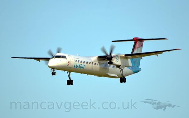 Side view of a white, high-winged, twin propellor-engined plane flying from right to left at a low altitude, with undercarriage fully extended from the rear of the engine nacelles, and flaps slightly extended from the rear of the wings, suggesting it is about to land.
There is a red lion standing on it's rear legs on the l;ower forward fuselage, just forward of the dopor, with blue "LuxAir" titles below the passenger cabin windows just aft of the door.
There is the blue outline of a circle containing a stylised blue flying bird on the sides of the engine nacelle.
The tail is red and the top, blue at the bottom, and has a white stripe in between these colours, and is textured to look like cloth, so that it resembles a waving flag - specifically, the flag of Luxembourg.
The same "flying bird in a circle" logo is at the top of the tail, except this time it is a white outline of a circle and a white bird.
Pale blue sky fills the rest of the frame.