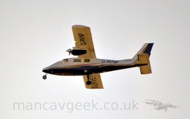 Slightly grainy side view of a white, high-winged, twin propellor-engined light aircraft flying from right to left ot a low altitude.
The plane has a dark blue belly, and red and yellow stripes running along the body.
The registration "G-RVNP" is visible under the left wing, and on the side of the rear fuselage.
The word "Survey" is visible under the right wing.
There are small, black "RavenAir" titles on the lower part of the tailfin.
Bright, hazy skies fill the rest of the frame.