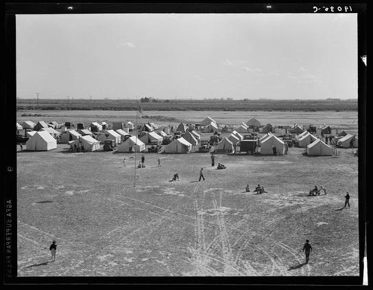 The image depicts an aerial view of several white tents arranged in rows on what appears to be a dry, flat landscape. These are likely part of a migrant labor camp situated in the Imperial Valley region of California during World War II, given its context as provided by Farm Security Administration (FSA) documentation from that time period.

Several individuals can be seen scattered around and interacting within this setting; some appear engaged in activities like playing baseball or lounging on the ground. The tents vary slightly in size but generally maintain a similar shape—rectangular with peaked roofs, typical of temporary housing used during wartime. In the background, there are sparse vegetation patches and distant structures that suggest an open rural environment.

The black-and-white quality of the photo indicates it was taken several decades ago, reflecting historical conditions for migrant laborers at such camps where families lived in tent cities often far from their permanent residences due to seasonal work requirements or displacement.