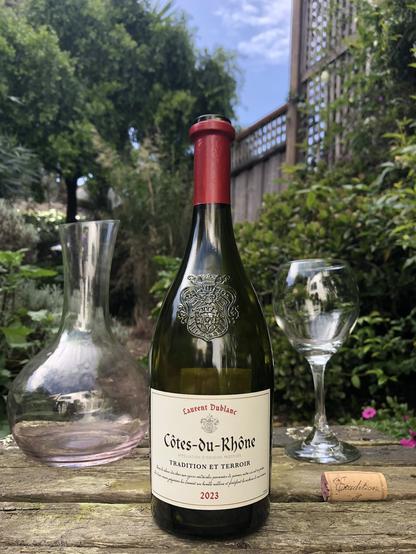 A color outdoor photo. A bottle of red wine in the center of a distressed wooden table in a garden. A wine decanter is in the background to the left, and a wine glass is to the right. A cork sits on the table in front of the glass. The text on the label, beginning at the top reads Laurent Dublanc. In the center of the label, Côtes du Rhône. Below that, Appellation d'origine protégée, followed by Tradition Et Terroir. At the bottom center, 2023. The green bottle is semi-transparent and includes a coat of arms embossed above the label.