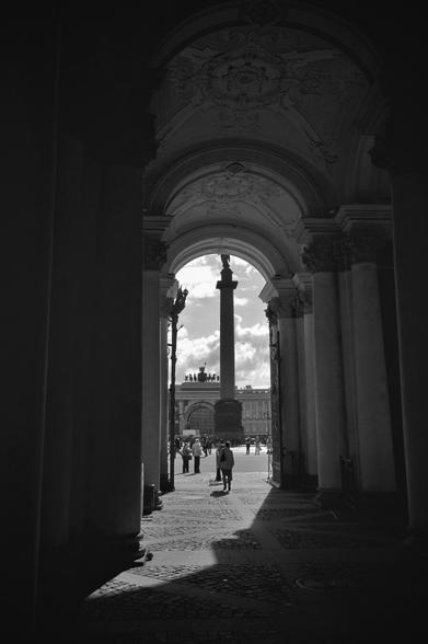 The image is a black and white portrait-orientated photograph taken from a dark, arched passageway looking out into a brightly lit plaza. The plaza is paved with cobblestones and features a tall, central column with a statue on top. Several people are visible in the plaza, walking in different directions. In the background, there is a long building with a colonnade and statues along its roofline. The sky above is visible with some clouds present. The foreground is dominated by the shadowed interior of the archway, showing stone pillars on either side and a decorated arched ceiling. Sunlight is casting shadows on the cobblestones in the plaza.