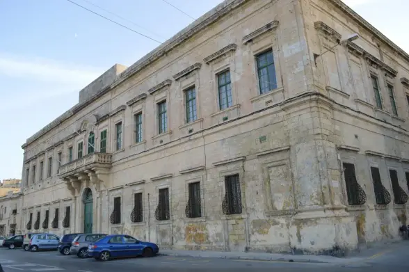The image shows a historical building, likely characterized by its aged architecture. It features multiple windows with decorative metalwork bars and a grand entrance. The walls appear to be made of stone, displaying a weathered appearance, which suggests the building has been around for quite some time. There are also some parked cars in the foreground.

Image Credits: Wikimedia / Continentaleurope / CC BY-SA 4.0
