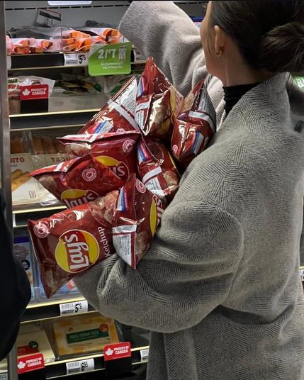 A woman in a gray beige wool coat digs in a convenience store shelf, arms overflowing with bags of Lay's Ketchup potato chips.