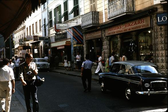 A crowded street scene with various shops: Bombay-Bazaar, Kodak, Remington Agency, Borsalino, etc.