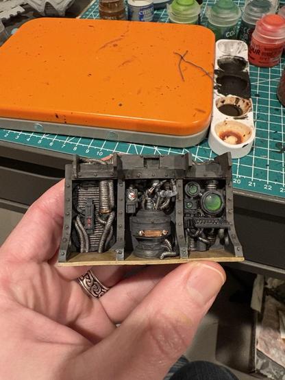 A wall panel in the engine room of the Reaver Titan. The bulkhead is a dirty gray. In the left rack position are exhaust vents and pipes. The center rack is a servitor in full on body horror mode, looking like a human battery from Matrix in a tank. On the right is a bank of screens and a terminal, a slight green color. The floor is a dirty tan. The whole thing has an oily / dirty vibe.