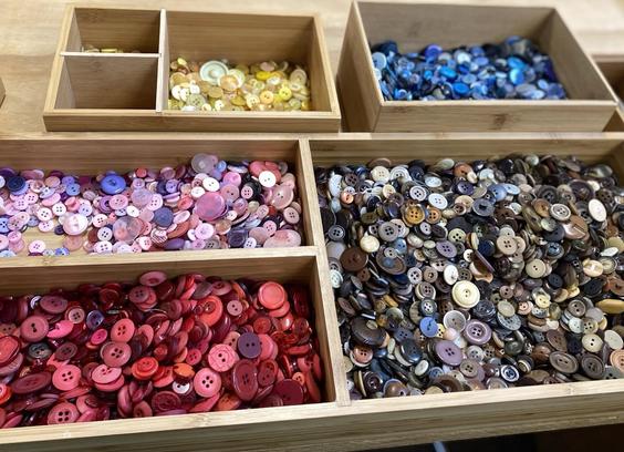 Several topped light wooden containers with buttons sorted into colours: yellow, blue, black/brown/grey/white, red, and pink/purple.