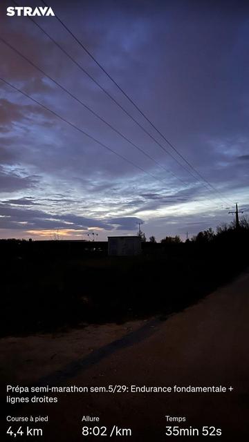 Point du jour par une météo couverte et venteuse sur un chemin de terre bordé d’arbres et de bâtiments industriels en contre-jour. Des statistiques Strava sont visibles.