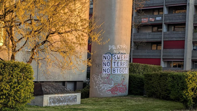Photo description: A blooming tree on the left in front of a building, balconies of another building on the right. In the middle a round concrete silo with a feminist collage on top that reads: NO SWERFS NO TERFS NO BIGOTS