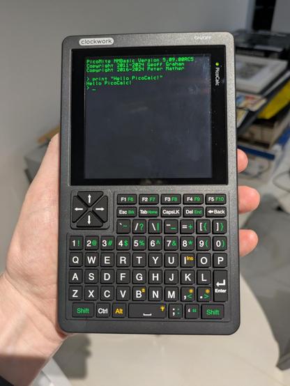 Photo of me holding the PicoCalc in my left hand. It's kind of like a large GameBoy with a QWERTY keyboard under the display. The display is showing a BASIC shell in green text. I have written: print "Hello PicoCal!", which it has echoed out on the next line.