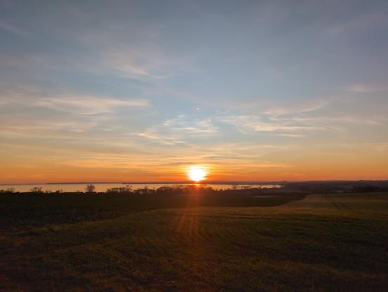 Das Bild zeigt eine atemberaubende Sonnenuntergangslandschaft. Der Himmel ist in warmen Farben wie Orange, Gelb und Rosa gefärbt, während die Sonne am Horizont untergeht. Die Sonne erzeugt einen leichten Glanz auf den Wolken und dem Wasser, das im Hintergrund zu sehen ist. Der Horizont ist von einem sanften Hügel bedeckt, der mit grünem Gras bedeckt ist. Im Vordergrund erstreckt sich ein Feld, das von der Sonne in ein sanftes Licht getaucht wird. Die Szene ist ruhig und friedlich, mit einer natürlichen Schönheit, die die Schönheit des Abendhimmels und der Landschaft hervorhebt.