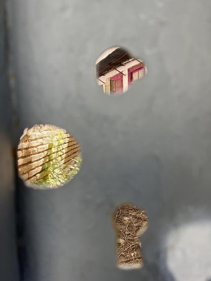 Behind a slab of metallic gate, a sunlit ruin is only visible through three holes in the metal. 
Bottom right, a disused keyhole shows dirt, brown twigs, leaf litter.
Middle height on the left, a round hole that may have once groused a door handle. Sunlit old style brick wall, with nettles.
Top right, it could be a keyhole again, circular with a small bump, two wooden windows painted red, with glass, one shutter open, a small corrugated metal awning above. The house is painted white and looks weathered but intact.