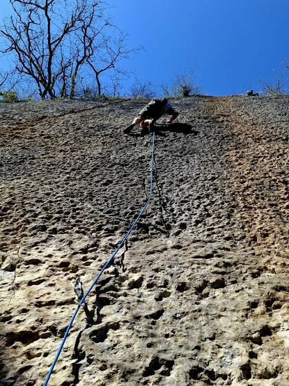 A person climbing the sunlit wall from the previous picture, seen from below. A rope attached to bolts via quickdraw leads up to the person.
The wall is full of small dimples and some trees and a blue sky can be seen at the top.