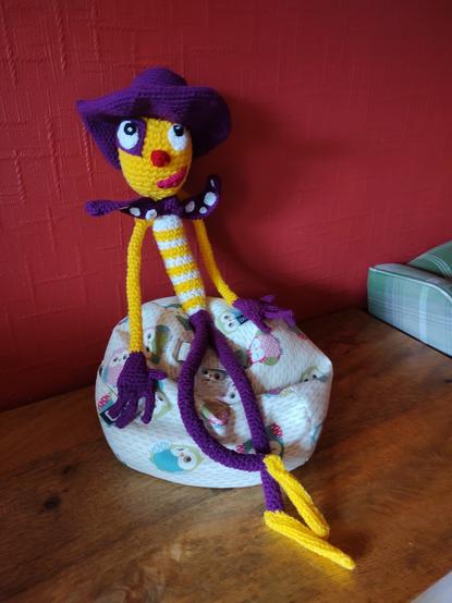 The crochet version of the long skinny chap in the first image. He is sitting on a little owl bean bag next to a red wall with one of his legs crossed over the other and his hands splayed at his sides. He is looking directly at the camera.